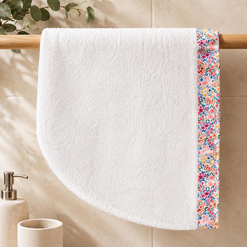 Puede incluir: Una toalla de mano blanca con una superficie de tela de rizo texturizada y un borde vertical decorado con un colorido estampado floral de peque&ntilde;as flores rosas, naranjas, amarillas y azules sobre un fondo blanco, colgada de una barra de madera.