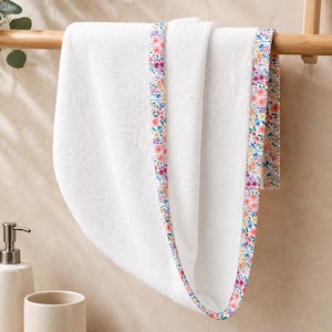 A white, fluffy baby towel with a floral patterned trim in pink, yellow, and blue. The towel is folded and hanging on a wooden rack, with greenery in the background. The towel is soft and absorbent.