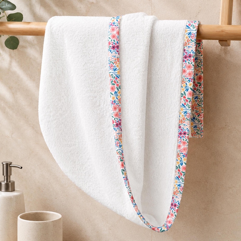 A white, fluffy baby towel with a floral patterned trim in pink, yellow, and blue. The towel is folded and hanging on a wooden rack, with greenery in the background. The towel is soft and absorbent.
