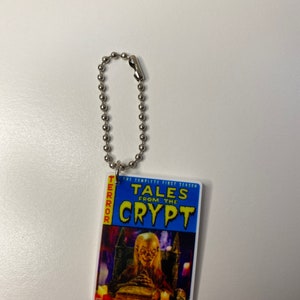 Puede incluir: Un llavero plateado con una portada de c&oacute;mic de Tales from the Crypt azul y amarilla. La portada muestra una figura espeluznante sentada en un trono con un libro en su regazo.