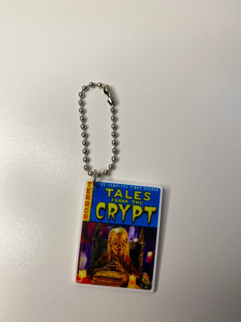 Puede incluir: Un llavero plateado con una portada de c&oacute;mic de Tales from the Crypt azul y amarilla. La portada muestra una figura espeluznante sentada en un trono con un libro en su regazo.