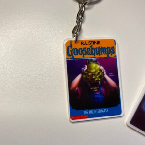 Puede incluir: Un llavero de Goosebumps con una portada de libro azul y naranja con el t&iacute;tulo "Goosebumps" y el texto "R.L. Stine" sobre el t&iacute;tulo. La portada del libro presenta una ilustraci&oacute;n de una persona con una m&aacute;scara de monstruo verde que le cubre la cara. El texto "The Haunted Mask" est&aacute; impreso debajo de la ilustraci&oacute;n. El texto "SCHOLASTIC" est&aacute; impreso en rojo en la parte inferior de la portada del libro.