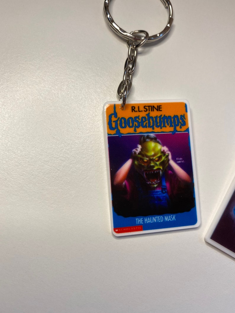 Puede incluir: Un llavero de Goosebumps con una portada de libro azul y naranja con el t&iacute;tulo "Goosebumps" y el texto "R.L. Stine" sobre el t&iacute;tulo. La portada del libro presenta una ilustraci&oacute;n de una persona con una m&aacute;scara de monstruo verde que le cubre la cara. El texto "The Haunted Mask" est&aacute; impreso debajo de la ilustraci&oacute;n. El texto "SCHOLASTIC" est&aacute; impreso en rojo en la parte inferior de la portada del libro.