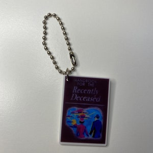 Puede incluir: Un llavero morado con una cadena plateada. El llavero presenta un peque&ntilde;o libro con el t&iacute;tulo "Handbook for the Recently Deceased". La portada del libro tiene una ilustraci&oacute;n colorida de dos personas caminando lejos de una puesta de sol.