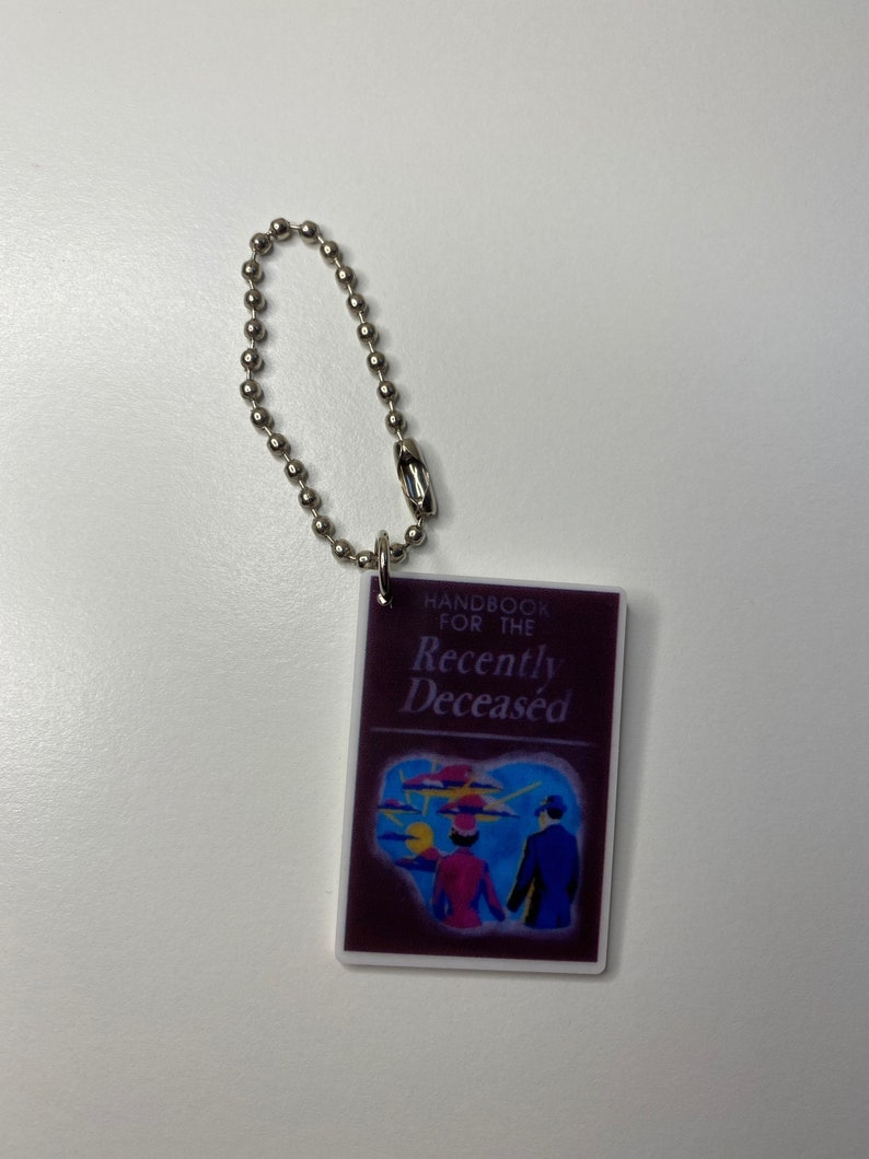 Puede incluir: Un llavero morado con una cadena plateada. El llavero presenta un peque&ntilde;o libro con el t&iacute;tulo "Handbook for the Recently Deceased". La portada del libro tiene una ilustraci&oacute;n colorida de dos personas caminando lejos de una puesta de sol.