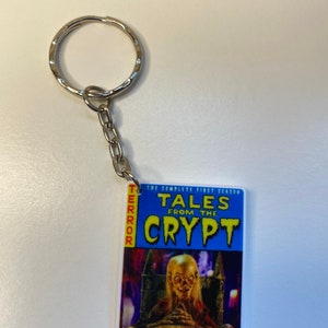 Puede incluir: Un llavero azul y blanco con una portada de c&oacute;mic para "Tales from the Crypt: The Complete First Season". La portada presenta una figura espeluznante, p&aacute;lida, con cabello largo sentada en un trono en un castillo oscuro.