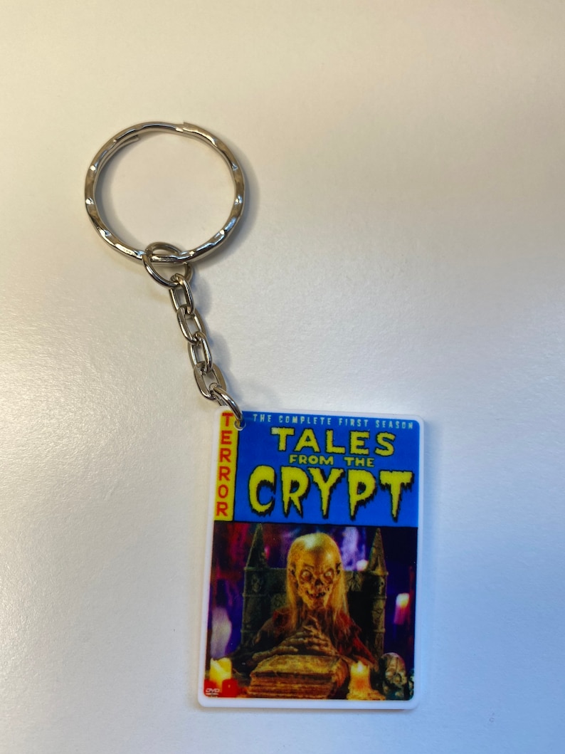 Puede incluir: Un llavero azul y blanco con una portada de c&oacute;mic para "Tales from the Crypt: The Complete First Season". La portada presenta una figura espeluznante, p&aacute;lida, con cabello largo sentada en un trono en un castillo oscuro.