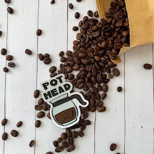 May include: A white sticker with the text "Pot Head" and a cartoon image of a coffee pot. The sticker is on a white wooden surface with coffee beans scattered around it.