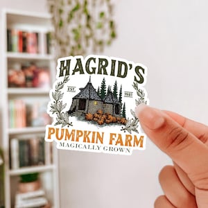 May include: A sticker featuring the text "Hagrid's Pumpkin Farm" with an illustration of a small house, pumpkins, and trees. The sticker has a vintage aesthetic with a white background and is held by a hand.