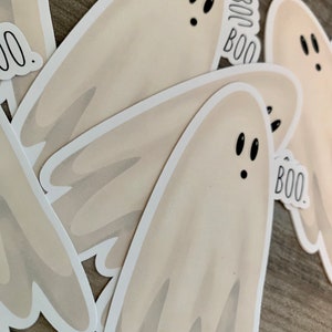 May include: A set of four white ghost stickers with black eyes and the word "BOO" written on them. The stickers are on a wooden surface.