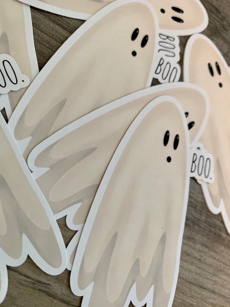 May include: A set of four white ghost stickers with black eyes and the word "BOO" written on them. The stickers are on a wooden surface.