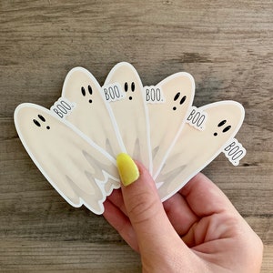 May include: Five white ghost stickers with black eyes and the word "BOO." printed on each one. The stickers are arranged in a fan shape and are being held by a hand.