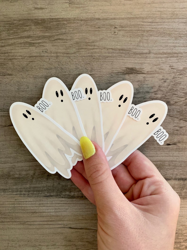 May include: Five white ghost stickers with black eyes and the word "BOO." printed on each one. The stickers are arranged in a fan shape and are being held by a hand.