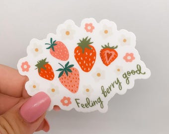 May include: A white sticker with a cluster of strawberries and small flowers. The strawberries are red and pink with green stems. The sticker has the phrase "Feeling berry good" in green script.