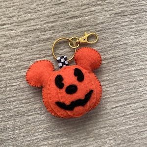 May include: An orange felt keychain shaped like a pumpkin with mouse-like ears. The pumpkin features black felt eyes, a nose, and a jagged mouth. A black and white checkered ribbon connects the felt piece to a gold-toned metal keyring and lobster clasp.
