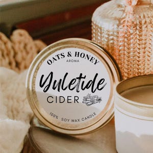 May include: A gold candle tin with a white label that reads "Oats & Honey Aroma Yuletide Cider 100% Soy Wax Candle". The label features a black and white illustration of cinnamon sticks and a star anise.