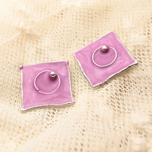 Peut inclure: Une paire de boucles d'oreilles carrées avec un anneau argenté et une petite perle argentée au centre. Les boucles d'oreilles sont de couleur violet clair.