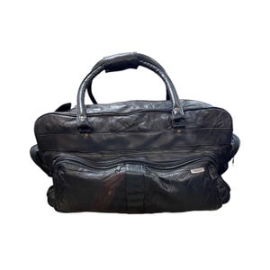 May include: Black leather duffel bag with a top handle and two smaller front pockets. The bag has a patchwork design and a zip closure. The handles are made of a sturdy leather material.