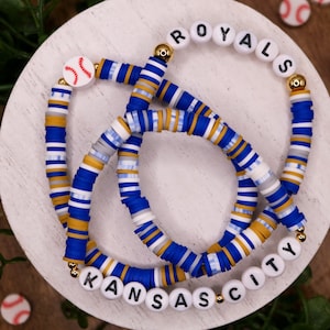 May include: Two layered bracelets with blue, white, and gold beads. The top bracelet spells "ROYALS" and the bottom bracelet spells "KANSAS CITY". A small red baseball charm is on the top bracelet.