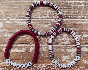 May include: Two red, white, and gray beaded bracelets with the words "Alabama" and "Roll Tide" spelled out in white beads.