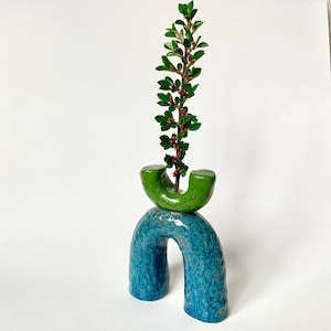 May include: A green ceramic vase with a unique arch shape, painted in a blue and green glaze. The vase is holding a sprig of green foliage with red berries.
