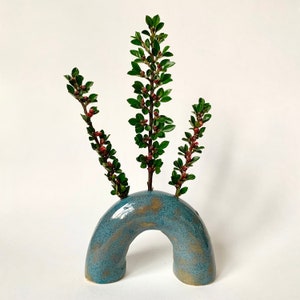 May include: A teal blue ceramic vase shaped like an arch with three sprigs of green foliage with red buds.
