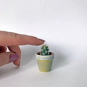 May include: A miniature green cactus with white spines in a decorative pot. The pot is painted with yellow vertical stripes and a gray and white checkered band. The pot is held by a finger with a purple nail.
