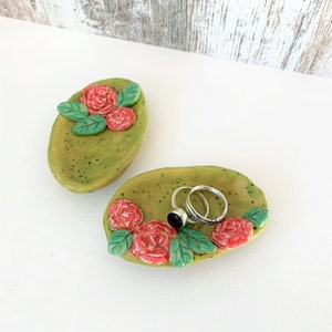 Könnte beinhalten: Zwei grüne Keramik-Ringteller mit roten Blumenakzenten. Der Teller rechts hat zwei silberne Ringe und einen Ring mit schwarzem Stein.