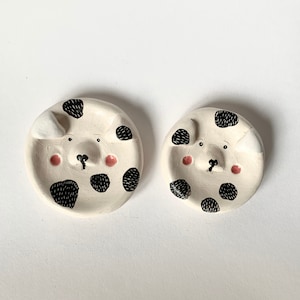 May include: Two white ceramic dishes with black and white polka dots and pink cheeks. Each dish features a simple black line drawing of a dog's face.