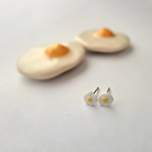 May include: A pair of white ceramic earrings shaped like fried eggs with yellow yolks. The earrings are on a white surface.