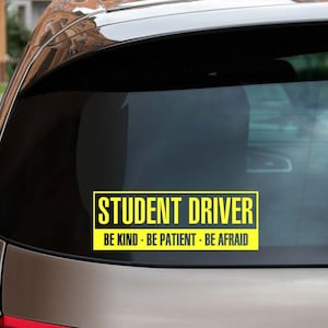 May include: A yellow and black student driver sign on the rear window of a car. The sign reads "Student Driver - Be Kind - Be Patient - Be Afraid".