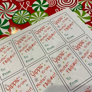 Puede incluir: Un conjunto de ocho etiquetas de regalo blancas con un diseño festivo de copo de nieve. Cada etiqueta presenta el texto "Yippie" y "Mother-F$@#er!" con la frase "Ki-Yay" repetida varias veces. Las etiquetas tienen espacios para escribir "Para:" y "De:".