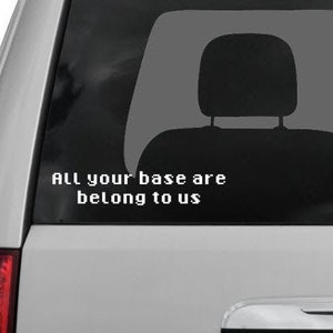 May include: A white car with a black rear window. The rear window has a white vinyl sticker with the text "All your base are belong to us".