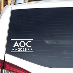 Puede incluir: Adhesivo para coche de vinilo blanco con el texto "AOC" encima de "2028" con dos estrellas en una ventana de coche. El adhesivo es rectangular y está diseñado para aplicarse en la ventana trasera de un vehículo.