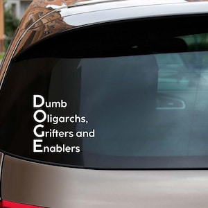 May include: A car window decal with white text that reads "Dumb Oligarchs, Grifters and Enablers." The decal is applied to the rear window of a brown car, with a side mirror visible.