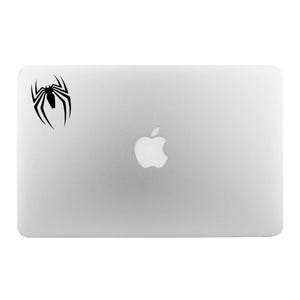 以下が含まれることがあります： 左上に黒いクモのデカール、中央にAppleのロゴが付いたシルバーのノートパソコン。クモのデザインは、8本の脚とセグメント化された体を持つ様式化されたシルエットです。