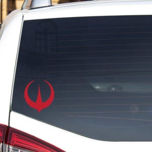 以下が含まれることがあります： 白い車のリアウィンドウに貼られた、赤いビニール製のステッカー。シンボルは、架空の組織またはフランチャイズを表す可能性のある、3つのポイントを持つデザインです。