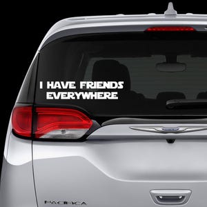 Pode incluir: Autocolante de vinil branco com o texto "I HAVE FRIENDS EVERYWHERE" no vidro traseiro de um carro prateado. O autocolante está centrado e em negrito, fonte sem serifa. As luzes traseiras do carro são vermelhas.