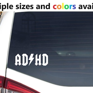 Puede incluir: Calcomanía de vinilo blanco para coche con el texto "AD⚡HD" en una fuente audaz y estilizada, que se asemeja a un logotipo de banda de rock. La calcomanía está aplicada a una ventana de coche. El texto "Multiple sizes and colors available" está en la parte superior.