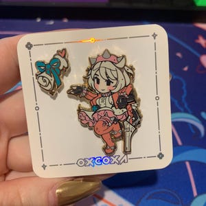 Könnte beinhalten: Zwei goldfarbene Emaille-Pins mit einer Comicfigur mit rosa Haaren und einem weißen Kleid, die eine Waffe hält. Die Figur steht auf einem weißen Hintergrund mit einem blauen und weißen Design. Die Pins befinden sich auf einer weißen Karte mit dem Text "oxcoxa" in blau und weiß.