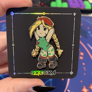 Op de afbeelding: Een gouden emaille pin met een cartoonfiguur met blond haar, een groene outfit en een rode hoed. Het personage staat met één hand op de heup en de andere arm omhoog. De pin is geplaatst op een zwarte achtergrond met een witte rand en de tekst "oxxcoxa" in een regenboogkleurige gradiënt.