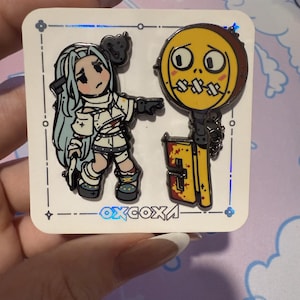 May include: Two enamel pins featuring a cartoon character with blue hair and a yellow key with a face. The key has a red and black design and the character is wearing a white dress with bandages. The pins are on a white background with a black border and the text "oxcoxa" in black.