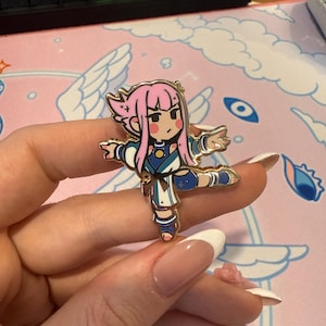 Pode incluir: Um broche de esmalte dourado com um personagem de desenho animado com cabelo rosa, um traje azul e branco e uma estrela no peito. O personagem está em uma pose de dança com uma perna levantada.