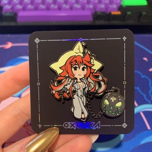 Peut inclure: Une épingle en émail noir et blanc représentant un personnage de dessin animé avec des cheveux roux, un chapeau jaune et une robe blanche. Le personnage tient une chaîne attachée à une citrouille noire et verte avec un visage effrayant. L'épingle est sur un fond noir avec une bordure argentée et le texte "oxgosa" dans une police holographique.