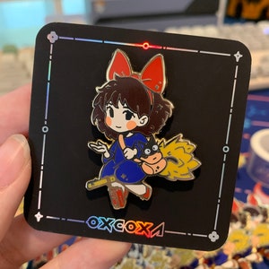 Puede incluir: Un pin de esmalte negro con un personaje de dibujos animados con un lazo rojo, un vestido azul y un gato negro. El personaje está volando en una escoba con una llama amarilla. El pin está sobre un fondo negro con un borde iridiscente arcoíris y el texto "oxcoxa".
