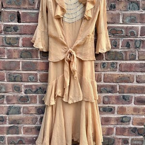 May include: A peach-colored vintage dress with a layered skirt and a matching top. The top features ruffled sleeves and a tie closure at the waist. The dress is hanging on a white hanger against a brick wall.