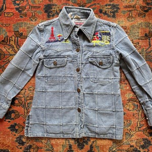 Puede incluir: Una camisa de mezclilla azul claro con un patrón de costuras en cuadrícula, cuello en punta y cierre frontal de botones. El área del pecho muestra parches bordados coloridos de la Torre Eiffel y un edificio con techo rojo y amarillo.