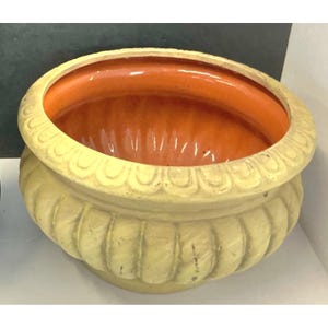 Puede incluir: Una maceta redonda de cerámica con un exterior texturizado de color amarillo pálido, detalles estriados verticales y un borde festoneado. El interior del cuenco tiene un acabado de esmalte naranja vibrante y liso que contrasta.