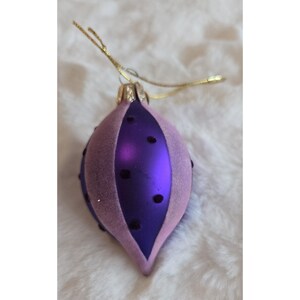 May include: A purple and pink Christmas ornament with a teardrop shape. The ornament has a shiny purple center with a pink, velvety texture on the sides. Small, dark dots are scattered across the surface, and a gold string is attached for hanging.