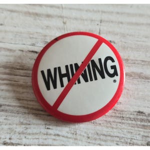 May include: A round pinback button featuring a white background with the word WHINING printed in bold black capital letters. A red circular border surrounds the text, and a red diagonal line crosses through the center of the button.